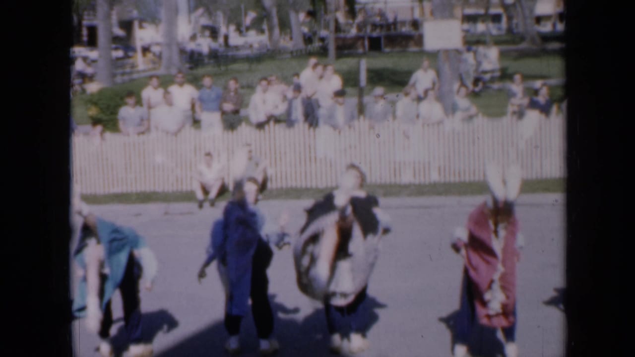 Dancers in traditional clothing turn and step across a paved area near spectators.