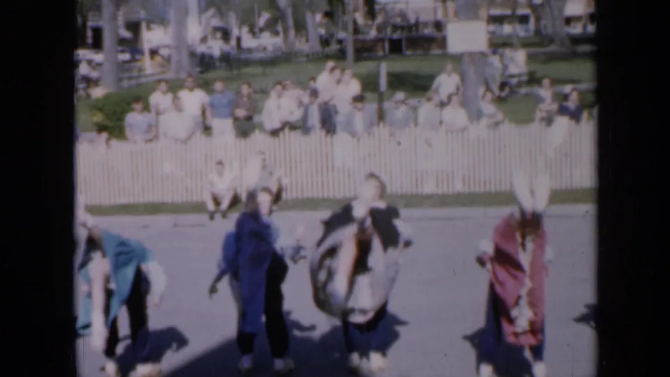 1959 Iowa park-festival frame with dancers in folk-style costumes moving across pavement as spectators sit beyond a white fence.