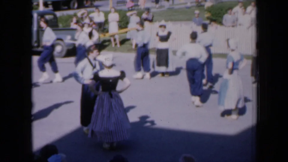 1959 Iowa 8mm frame showing a wider folk dance formation, a parked car, and spectators watching from behind a white fence.