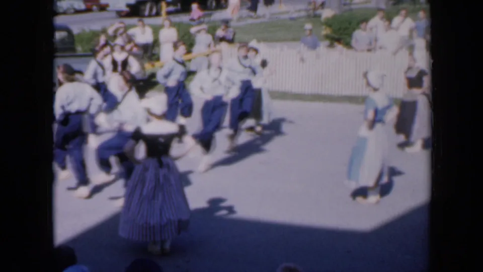 1959 Iowa home-movie frame of a folk dance group in motion, with full skirts, bonnets, spectators, and strong afternoon shadows.