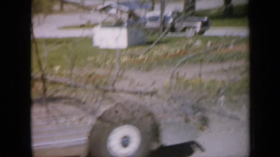 1959 Iowa home-movie frame with motion blur as a silver bus-like vehicle passes in front of the yard and garden path.