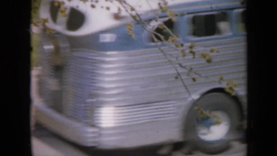 1959 Iowa frame showing the side of a silver bus-like vehicle moving past the residential yard, partly obscured by branches.
