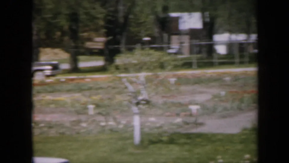 1959 Iowa frame with garden beds, a small tree, lawn, a parked mid-century car, roadside trees, and buildings beyond the fence.