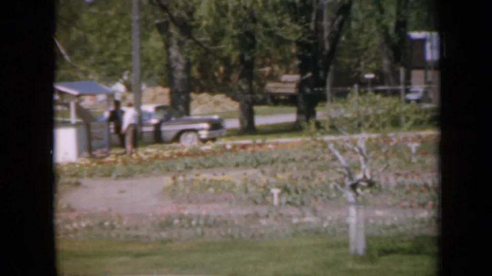 1959 Iowa home-movie view of two distant people near a parked car beside garden beds, trees, fencing, and a small roadside structure.