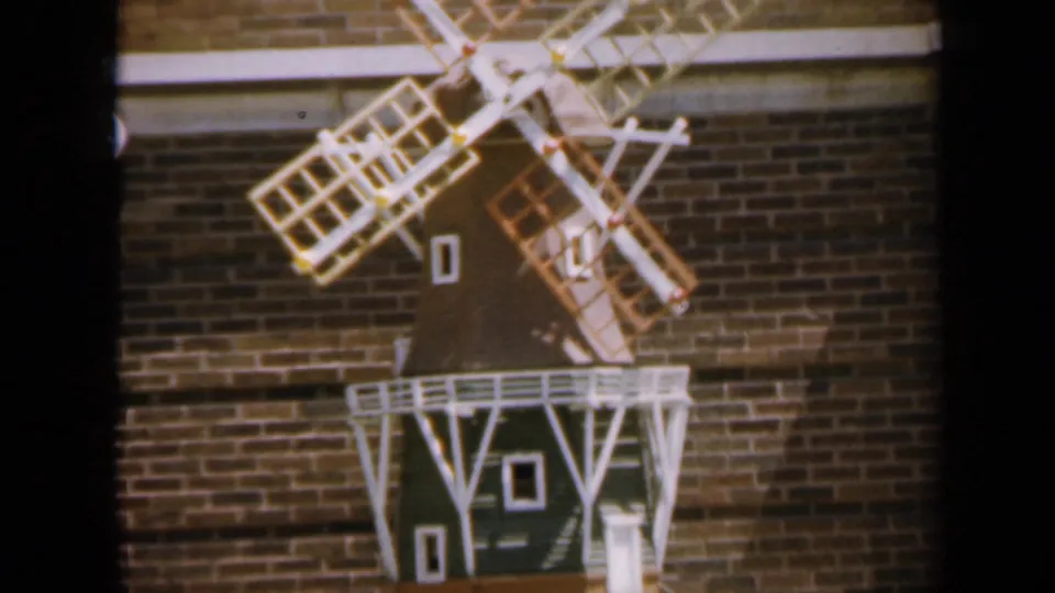 1959 Iowa frame showing a decorative windmill model mounted on a brick wall, with pale railings, lattice blades, and small painted windows.