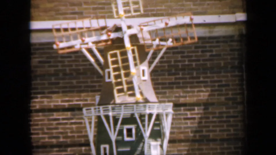 1959 Iowa 8mm frame showing a small decorative windmill with white railings against a brick wall.