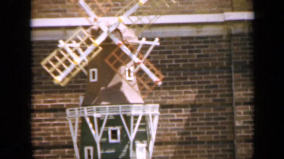 1959 Iowa home-movie frame of a small windmill with pale blades and white railings set against brickwork.