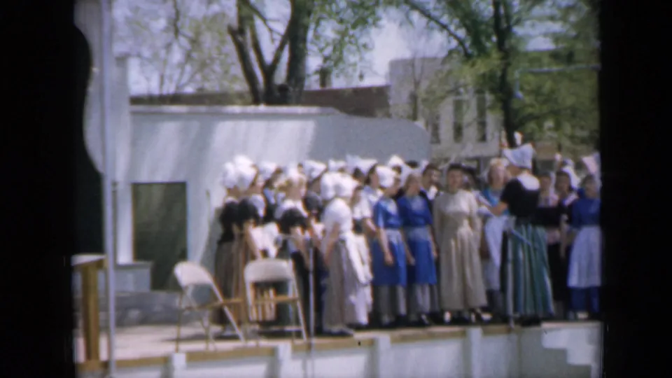 1959 Iowa home-movie frame showing a stage group, mostly women in bonnets and long dresses, outdoors near trees and a white structure.