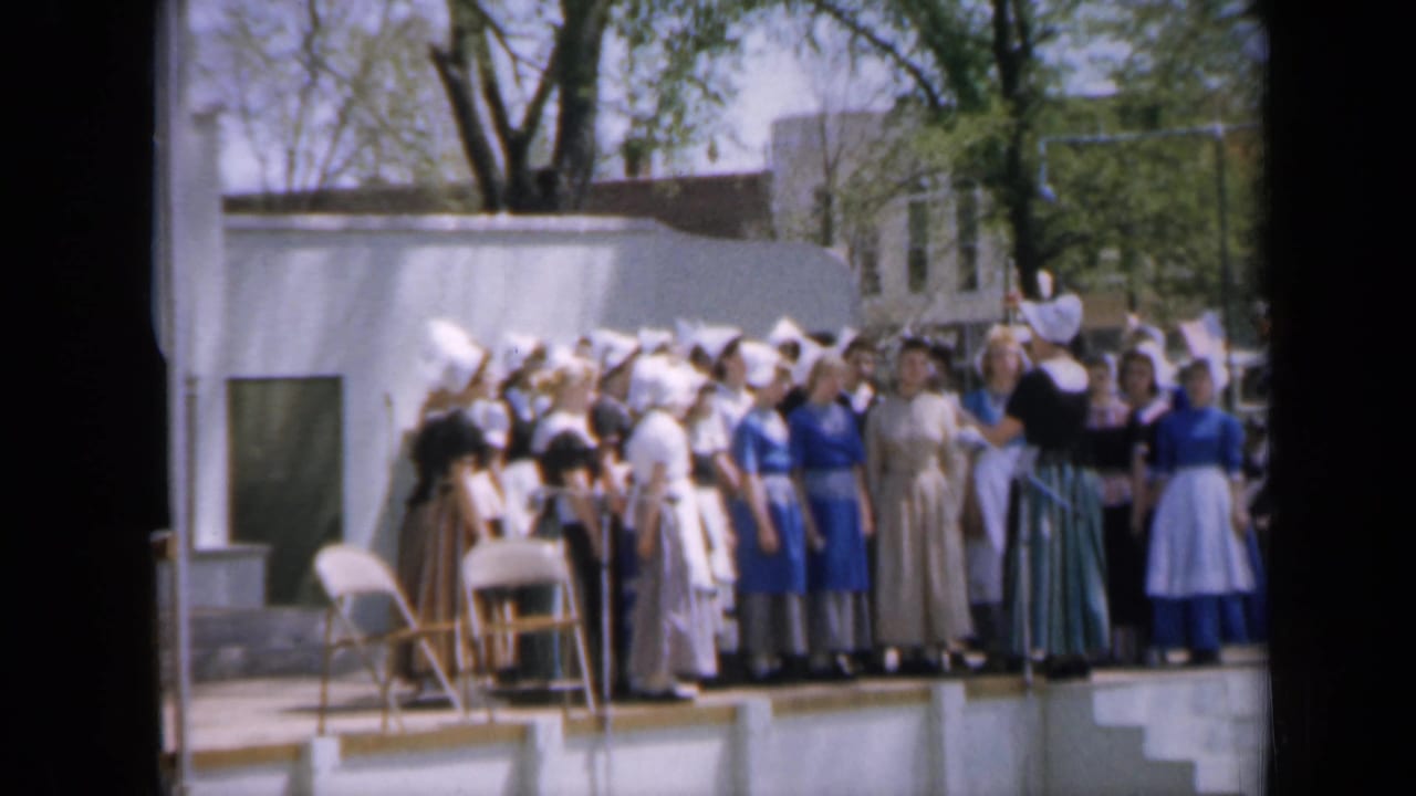 Costumed group stands on an outdoor stage with bonnets, long dresses, and folding chairs nearby.