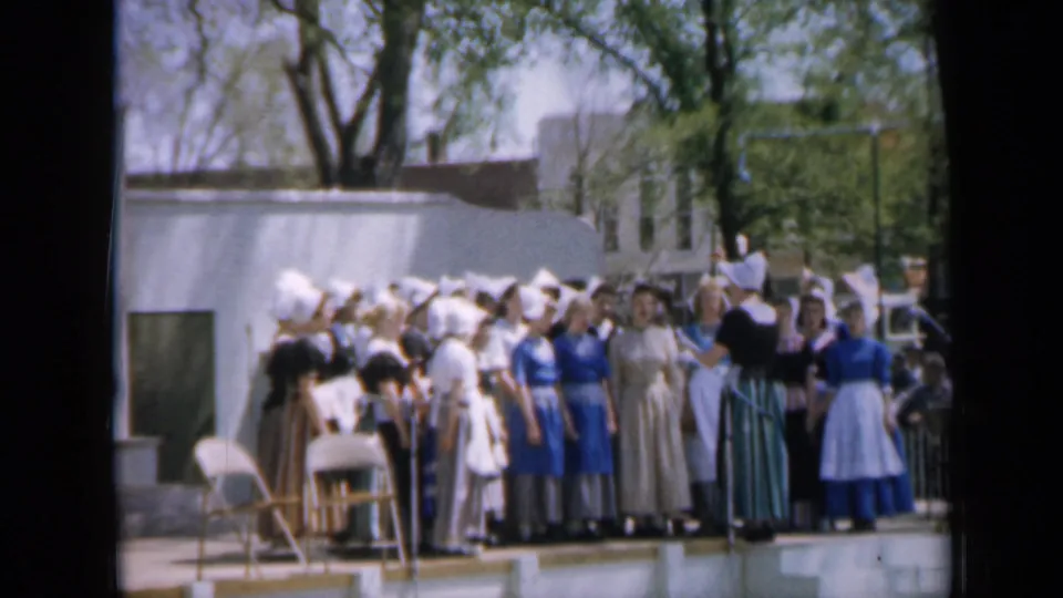 1959 Iowa 8mm frame of performers in bonneted period-style costumes grouped on an outdoor stage near folding chairs.