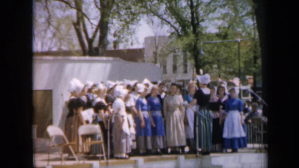 1959 Iowa frame showing a costumed girls' choir or pageant group on a small outdoor stage with trees and buildings behind.