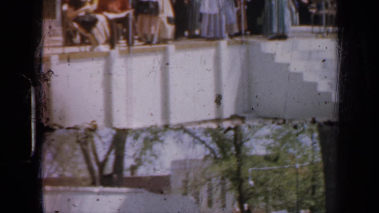 Guests in suits and long dresses stand on a white outdoor balcony above trees.