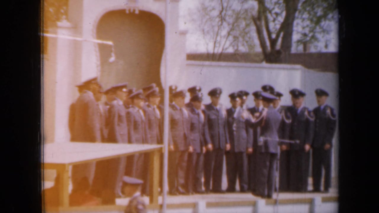 Uniformed personnel stand in formation beside a white arched structure during an outdoor ceremony.