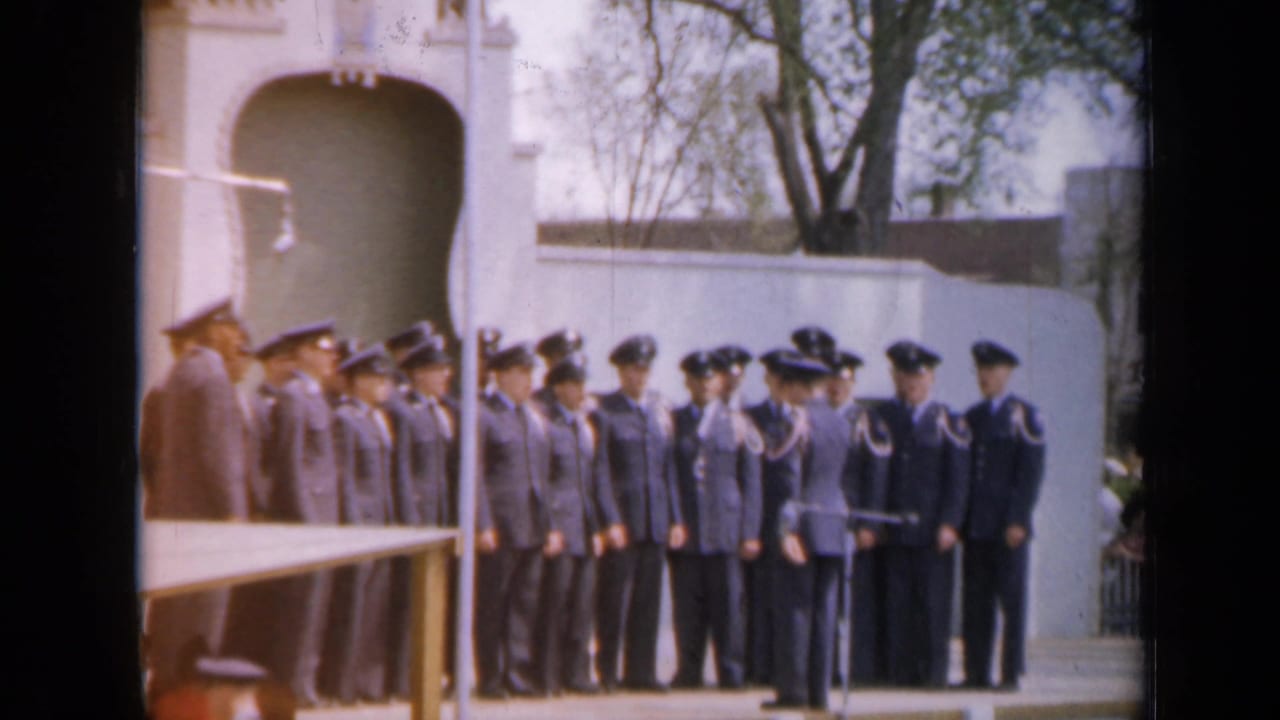Cadets in dark uniforms stand outdoors while an officer addresses them near an arched white building.