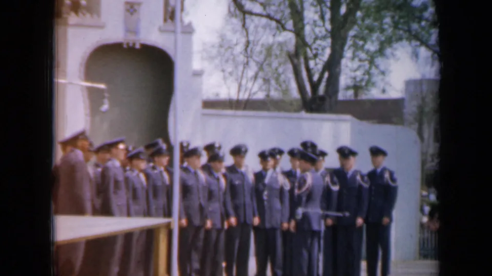 Iowa 1959 footage shows cadets in dark uniforms receiving baton-led instruction outside a pale building with trees behind it.