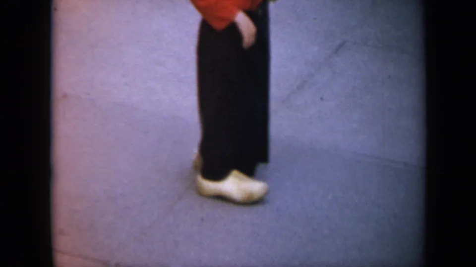 In the 1959 Iowa home-movie clip, a passerby in dark clothing, a red top, and white wooden clogs crosses close to the lens.