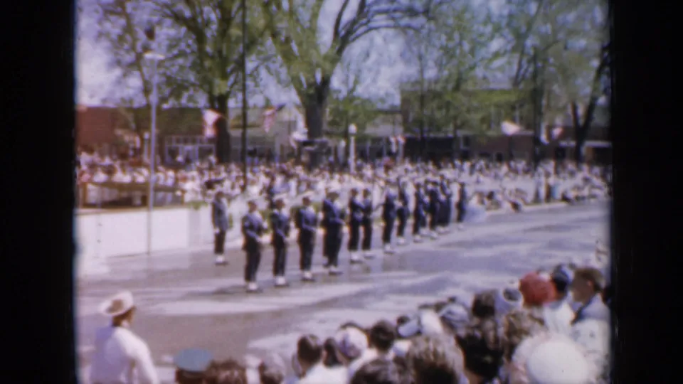 A 1959 Iowa parade scene shows uniformed soldiers lined up with rifles as spectators watch from a sunlit small-town street.