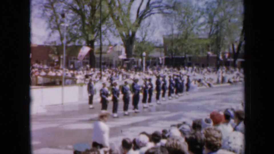 The Iowa home-movie frame shows a uniformed formation in a patriotic parade, with spectators, flags, trees, and mid-century buildings in view.
