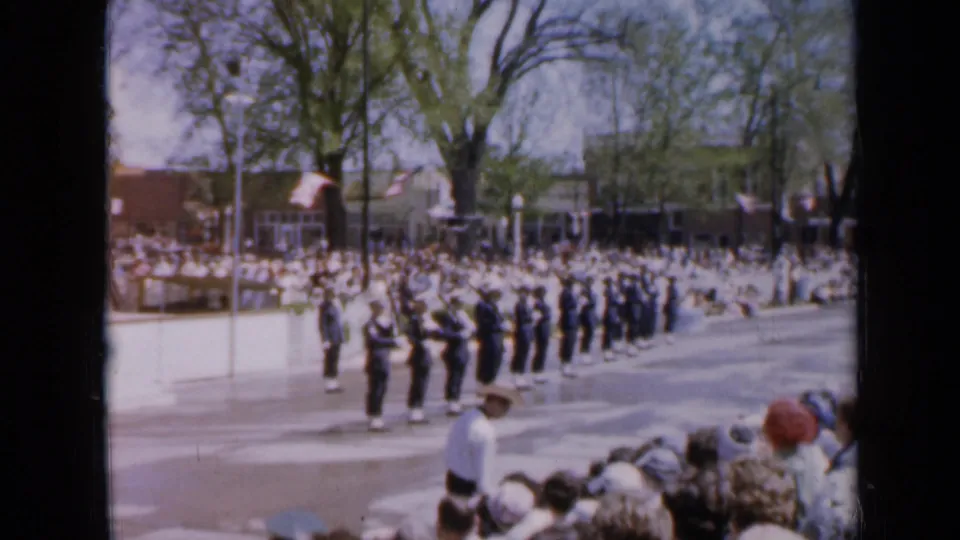 Uniformed soldiers with rifles stand in formation during the 1959 Iowa parade, watched by a curbside crowd under trees and American flags.