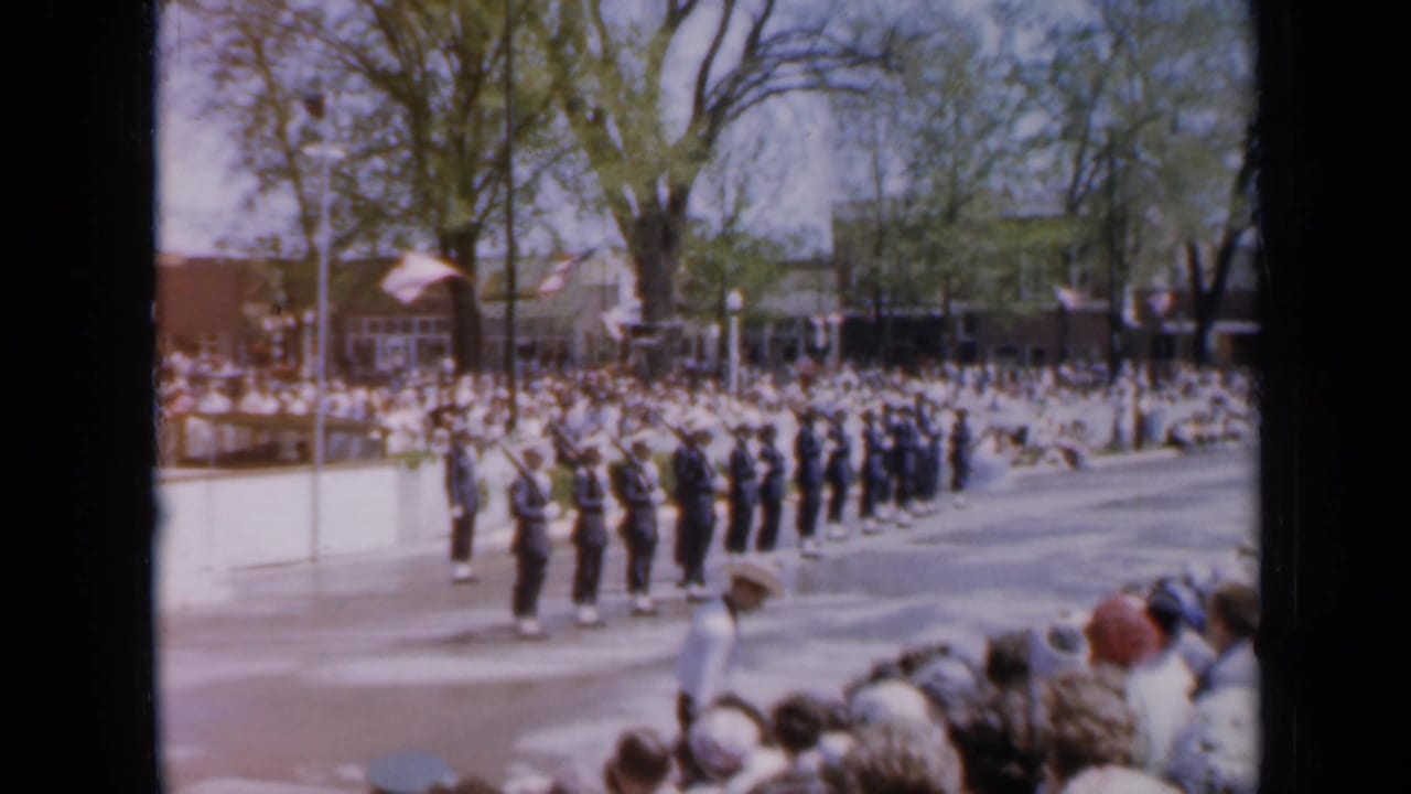 Uniformed marchers pass spectators along a sunny tree-lined street.