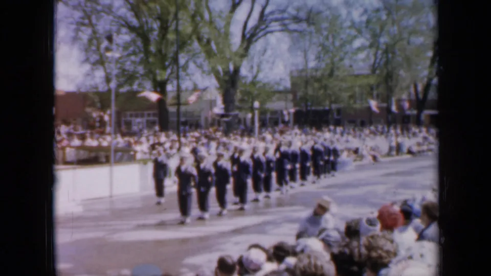 1959 Iowa parade view with uniformed marchers moving along the street as a dense sidewalk crowd watches.
