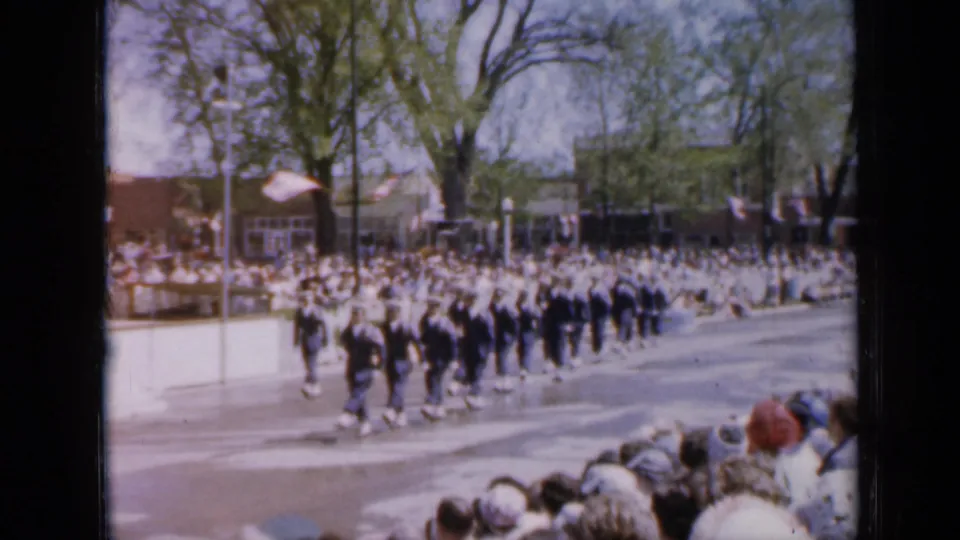 1959 Iowa home-movie frame of uniformed participants marching past spectators, trees, flags, and storefronts.