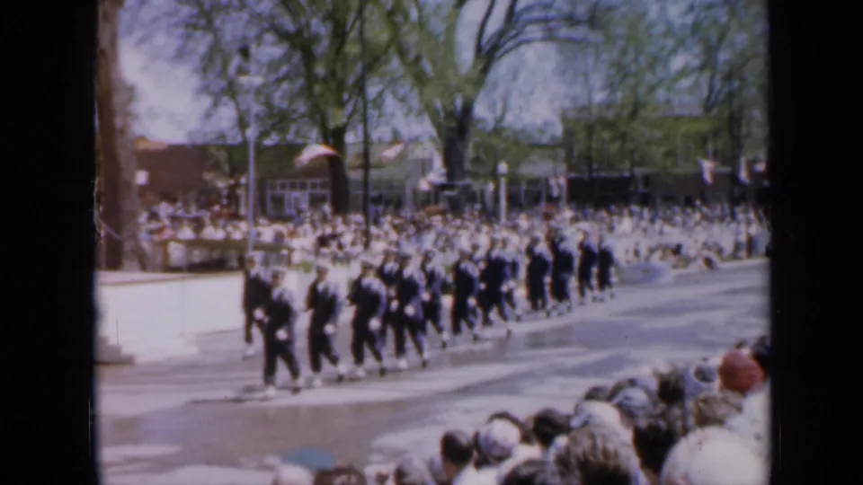 1959 Iowa parade frame showing dark-uniformed marchers passing a packed sidewalk crowd under tall trees.