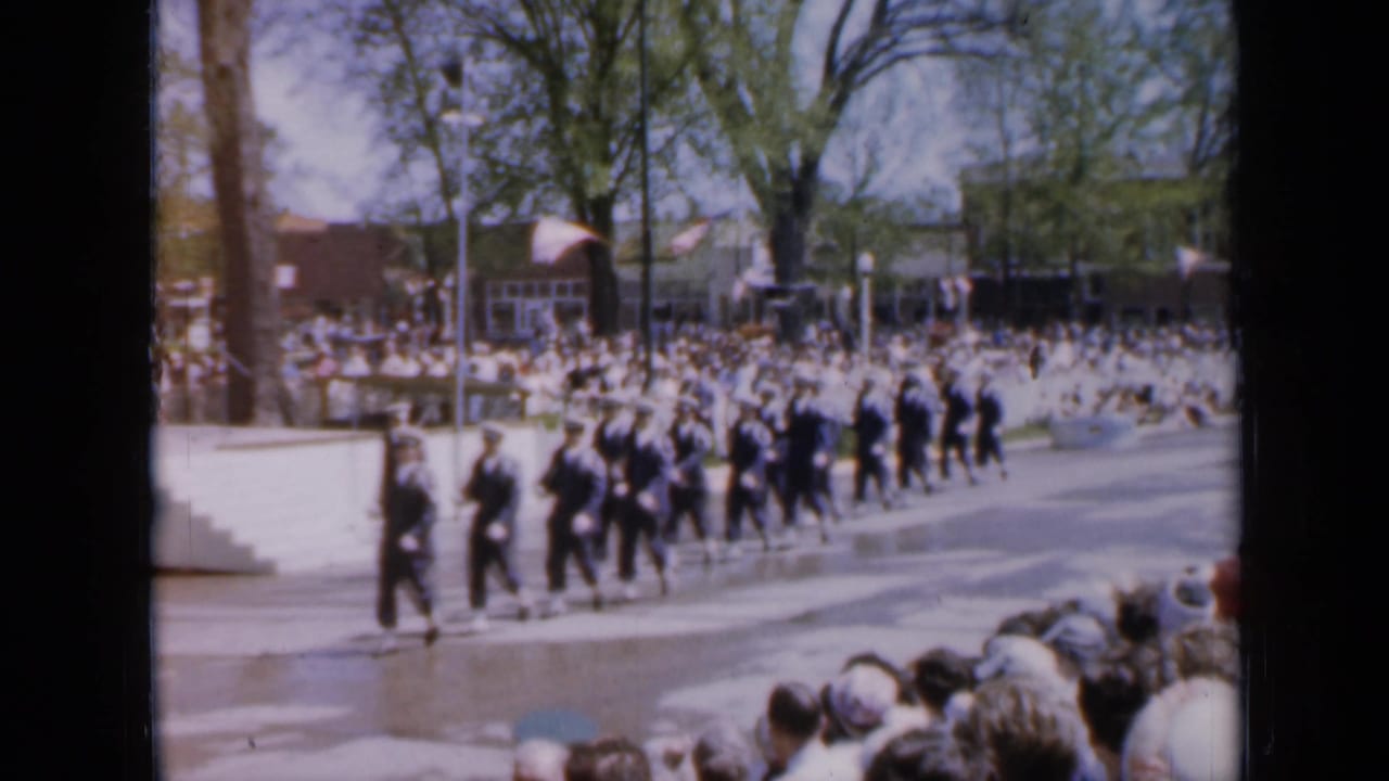 Uniformed soldiers march past spectators along a tree-lined street.