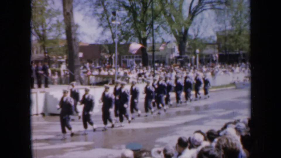 1959 Iowa parade view with uniformed soldiers moving along the street while a large public audience watches behind a low wall.