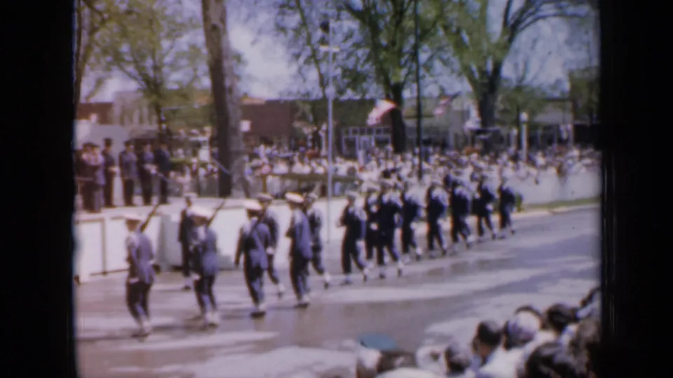 1959 Iowa home-movie frame showing uniformed marchers, some carrying rifles, passing spectators along a sunny street.