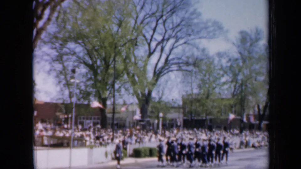 Wide 1959 Iowa parade frame with soldiers in formation, a large audience, flags, trees, and buildings lining the route.