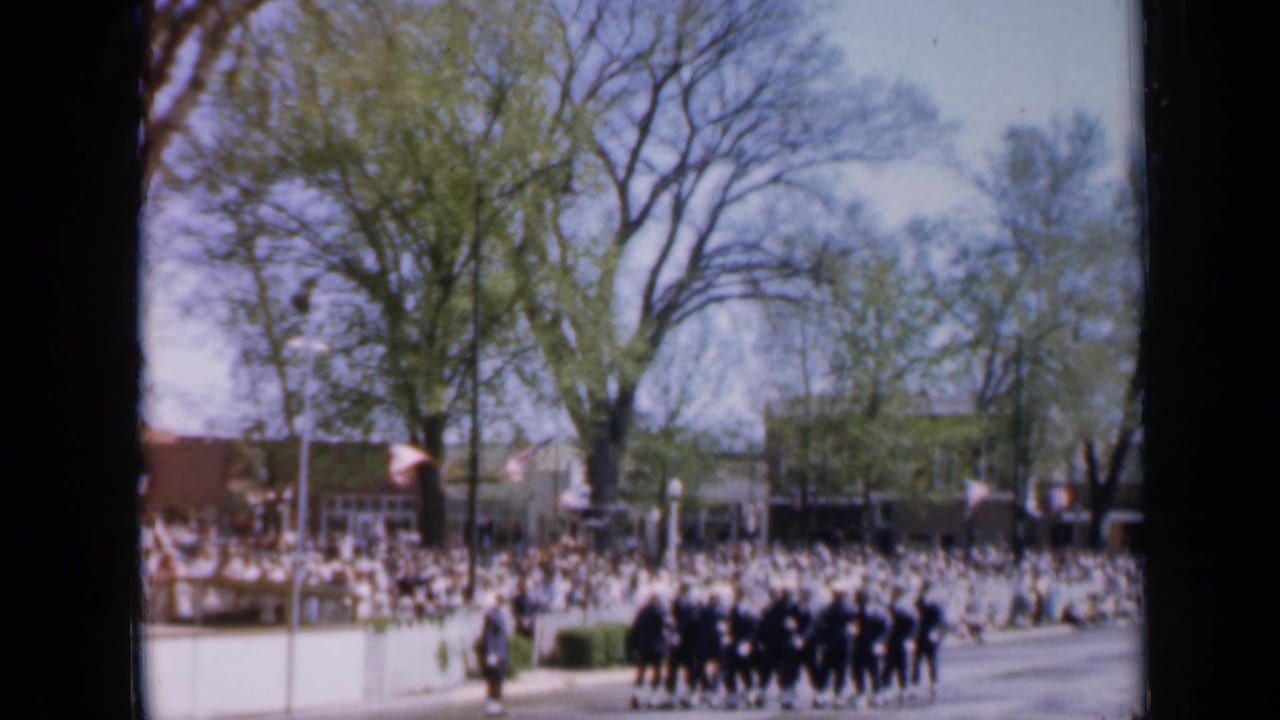 Uniformed marchers pass a large crowd on a tree-lined street.