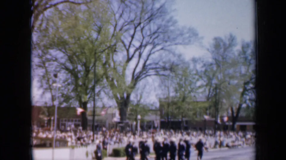 Soft 8mm frame from Iowa in 1959 showing uniformed parade participants before a crowded sidewalk.