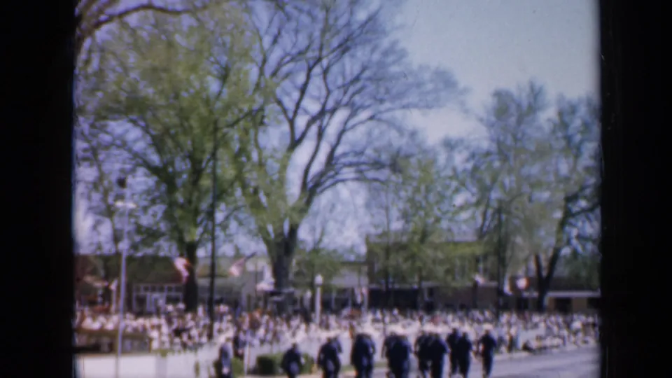 1959 Iowa parade frame with uniformed marchers near the foreground and spectators lining the opposite side of the road.