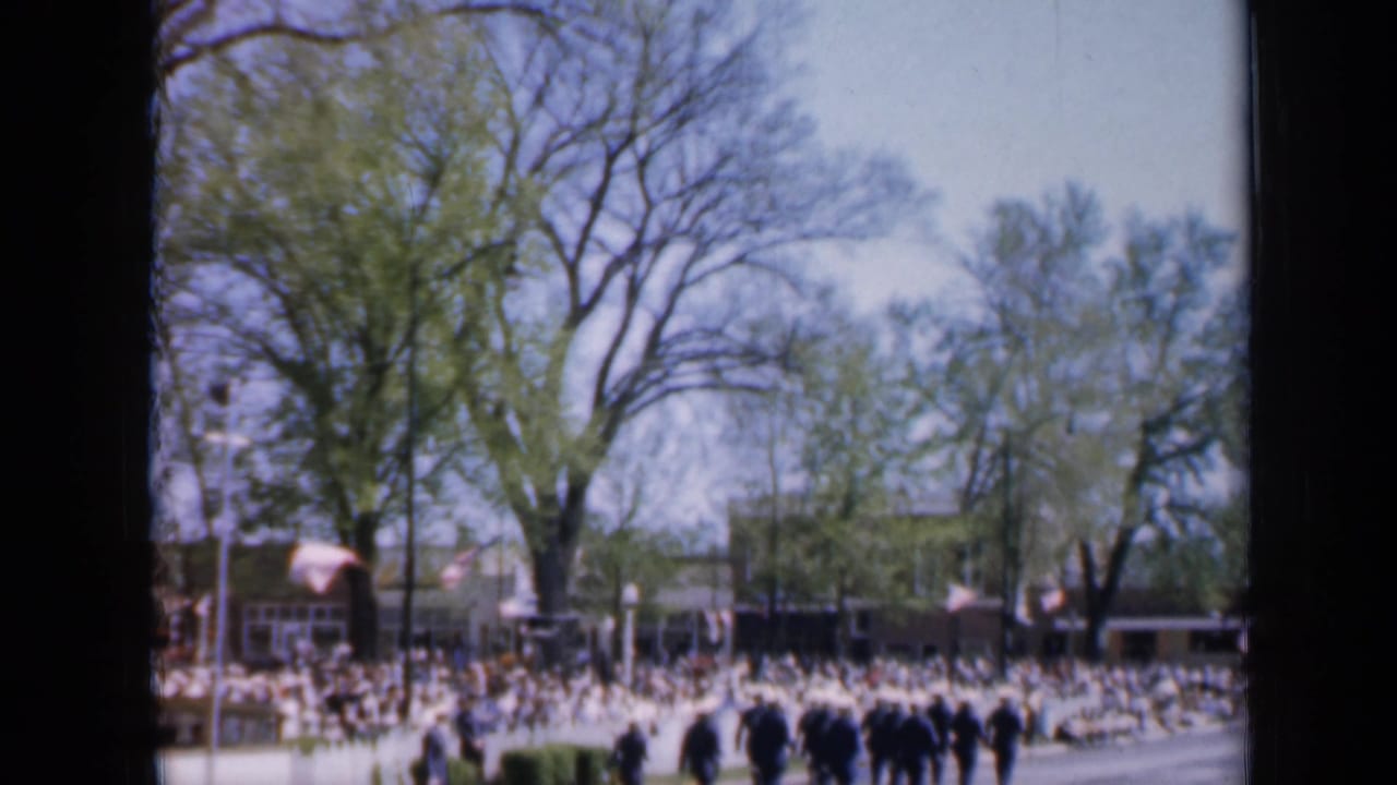 Wide parade route with marching uniformed men, spectators, trees, and flags.