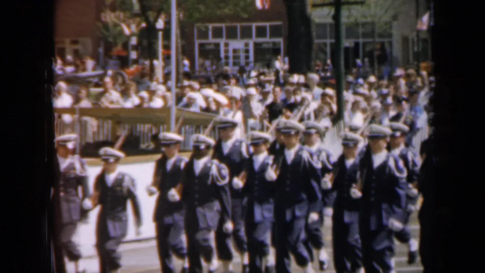 Uniformed marchers in white caps pass the camera during a 1959 Iowa parade, with spectators seated and standing behind the route.