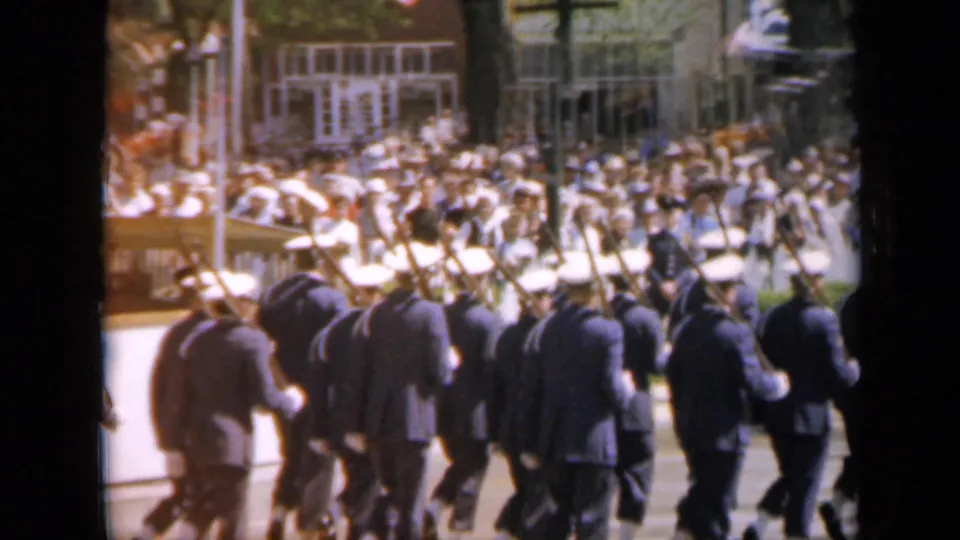 Rear view of a marching unit in dark uniforms during a 1959 Iowa parade, with spectators and building windows across the street.