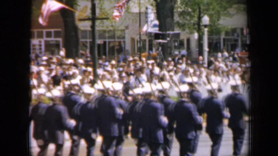 A 1959 Iowa parade frame shows the uniformed group from behind, American flags overhead, and spectators watching near storefront windows.
