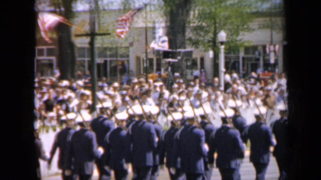 Uniformed men march with rifles past a sidewalk crowd on a sunny street.