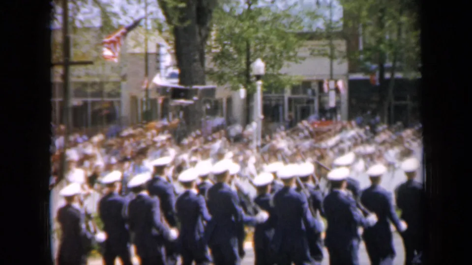 1959 Iowa parade frame with uniformed marchers carrying rifles, American flags overhead, and a large crowd along storefronts.