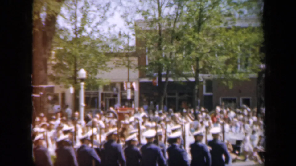 1959 Iowa home-movie view of uniformed men marching with rifles while spectators fill the sidewalk near downtown storefronts.
