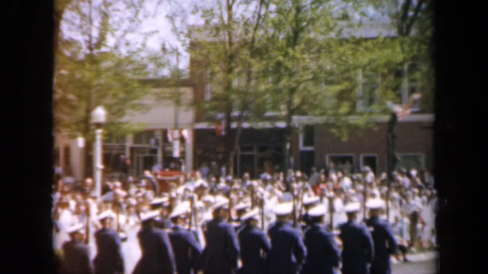 1959 Iowa street-parade frame showing men in dark uniforms and white caps marching with rifles in front of a dense sidewalk crowd.