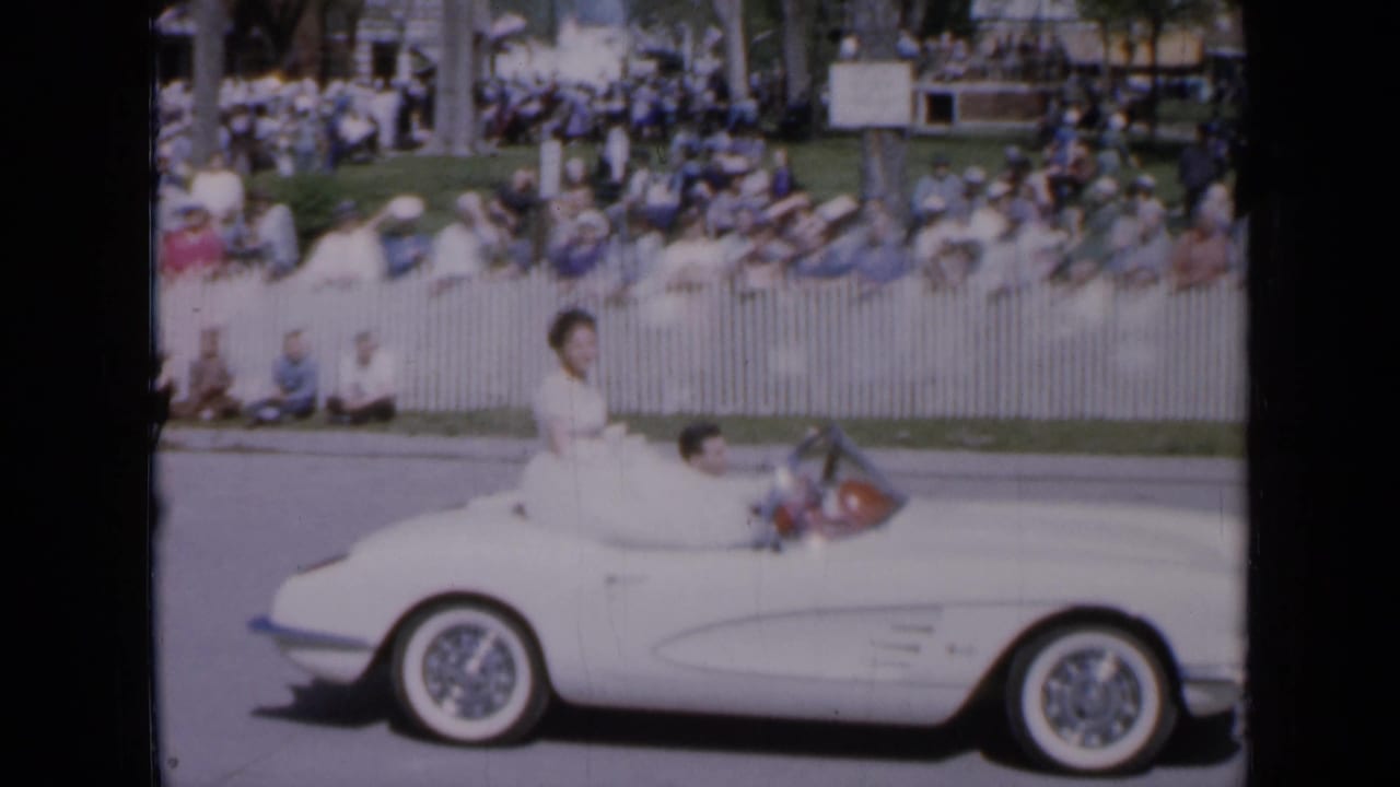 White convertible passes spectators as a woman in a white dress rides above the passenger area.