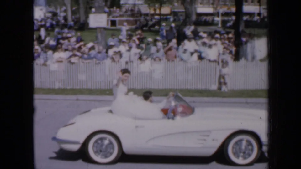The white parade convertible moves along the Iowa street as the woman in white faces the crowd and spectators watch from behind fencing.