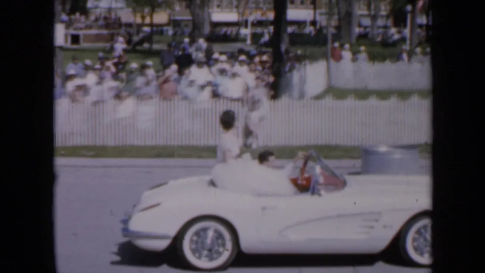 The home-movie camera tracks the rear of the white convertible while the woman in white passes the parade spectators in bright sun.