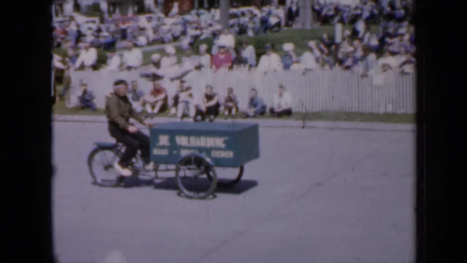 A man on a bicycle pulls a green vendor-style cart marked DE VOLHARDING along the parade route in the 1959 Iowa footage.