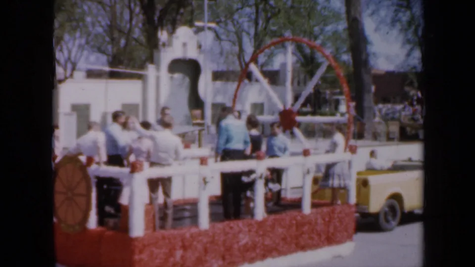1959 Iowa parade frame with people on a white-railed float and part of a light-colored vintage car at the curb.