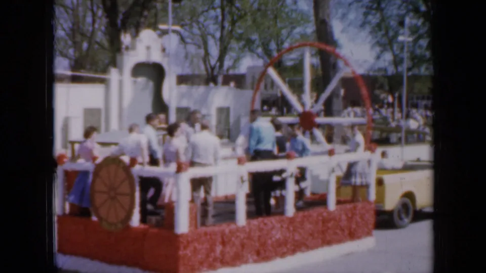 1959 Iowa home-movie frame showing a decorated float with wagon-wheel detail, people aboard, and spectators in the background.