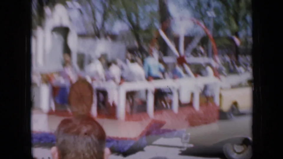 Soft 1959 Iowa frame of a parade float with people, patriotic-colored trim, and a light-colored car in the foreground.