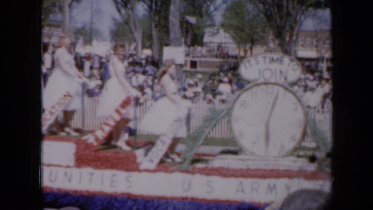 Women in white dresses ride a patriotic U.S. Army parade float with a large clock prop.